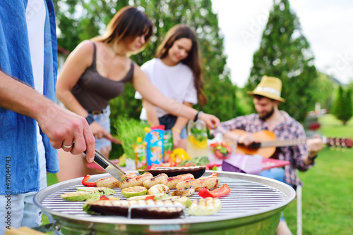 a man s hand holds a barbecue tongs with a juicy delicious meat steak against the background of a barbecue grill with meat and vegetables and a group of friends on a picnic who are having fun and