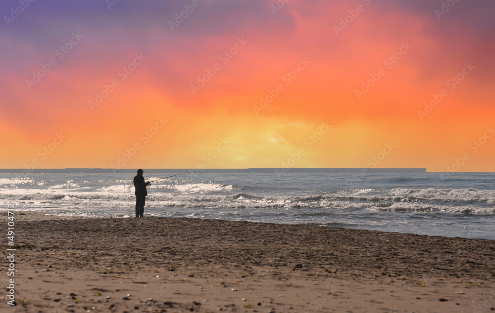 Fisherman is fishing on the seashore. Silhouette of a man with a fishing rod while fishing in sea on sunset. Fisher on coastline at sea with waves during storm and wind. Wave at rising storm in ocean.