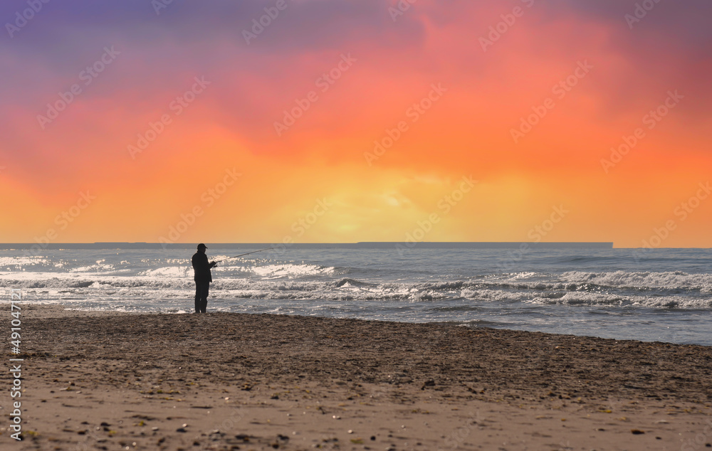 Fisherman is fishing on the seashore. Silhouette of a man with a fishing rod while fishing in sea on sunset. Fisher on coastline at sea with waves during storm and wind. Wave at rising storm in ocean. Fisherman is fishing on the seashore. Silhouette of a man with a fishing rod while fishing in sea on sunset. Fisher on coastline at sea with waves during storm and wind. Wave at rising storm in ocean.