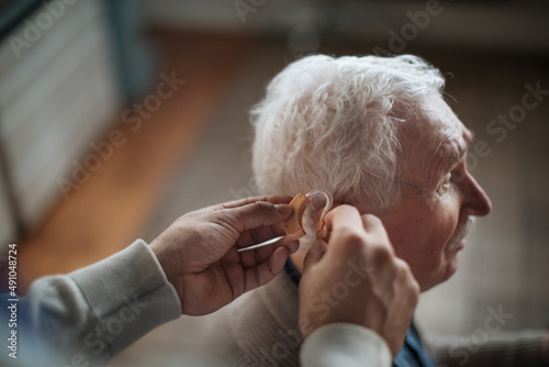 Close-up of caregiver man's hand inserting hearing aid in senior's man ear.