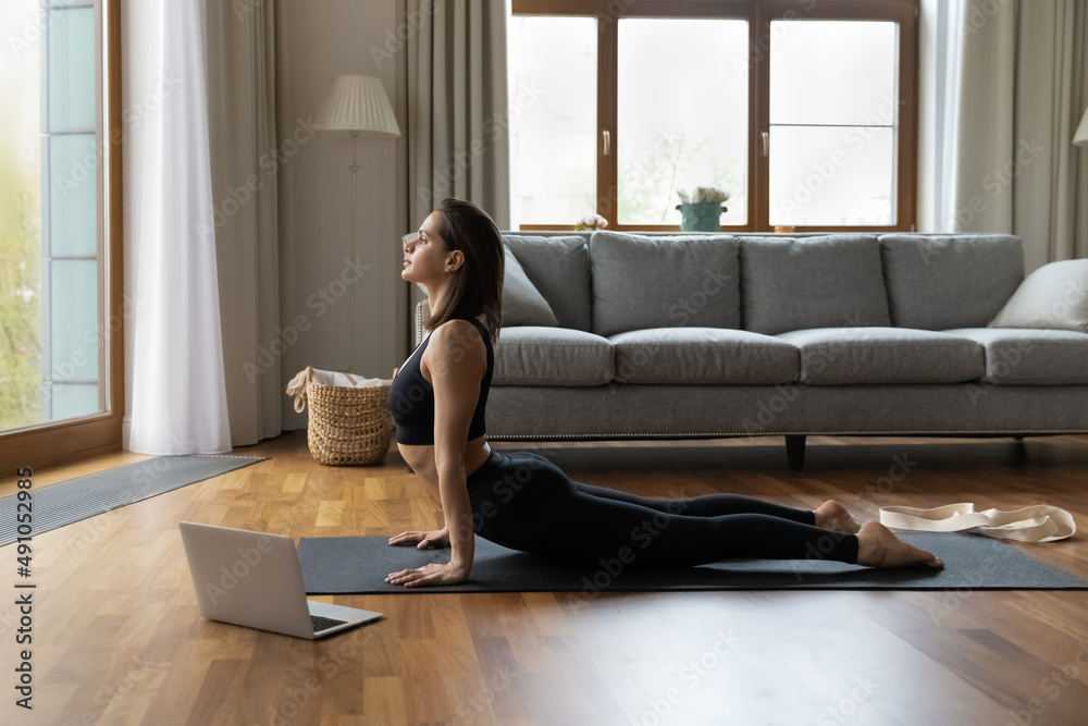 Distance yoga class, online training, work out at home in quarantine concept. Woman in black sportswear do stretching exercise, perform Upward Facing Dog asana on mat in living room near opened laptop Distance yoga class, online training, work out at home in quarantine concept. Woman in black sportswear do stretching exercise, perform Upward Facing Dog asana on mat in living room near opened laptop