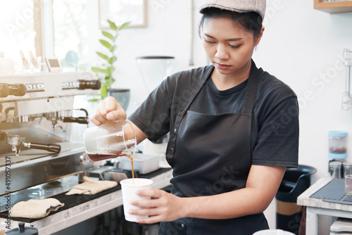 Smiling Asian barista young woman pouring hot black coffee into paper cup for according to the customer's order at counter bar in coffee shop.
