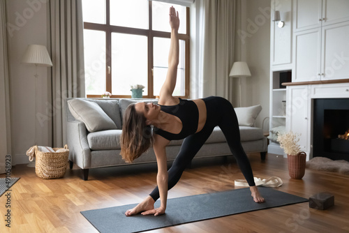 Young beautiful sporty woman practicing yoga doing Utthita Trikonasana exercise or Extended Triangle asana, working out wearing comfy black sportswear indoor. Sport at home, active lifestyle concept