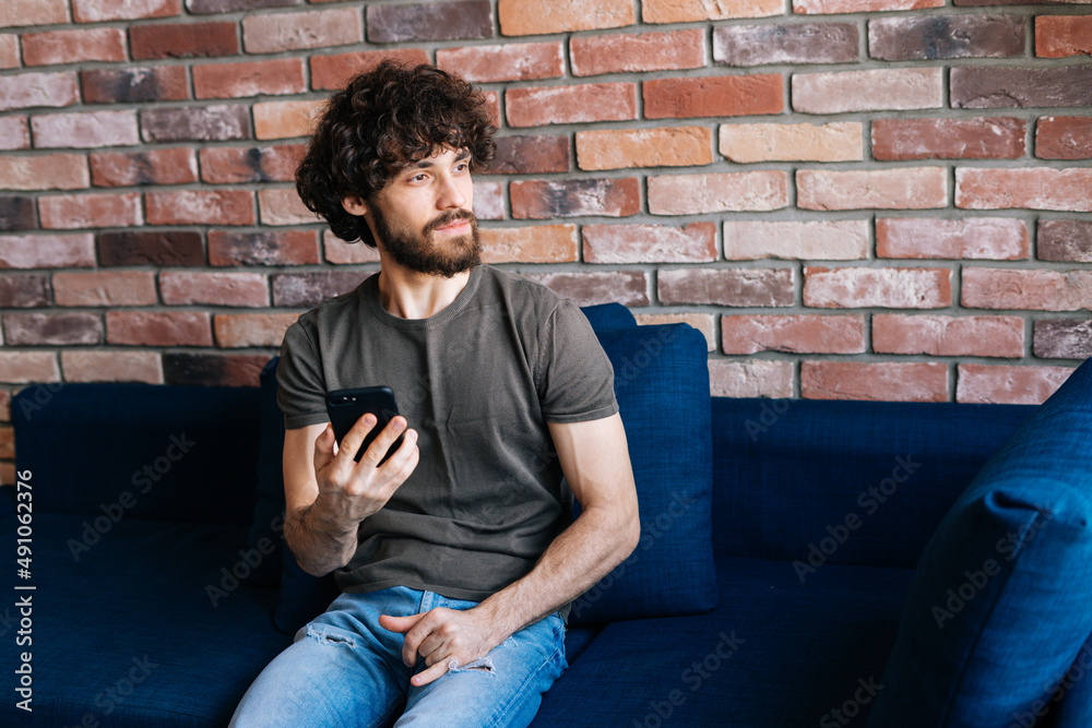 Portrait of thoughtful bearded young man looking away browsing wireless internet on mobile phone sitting on sofa at home. Confused male working, learning, blogging, chatting online.