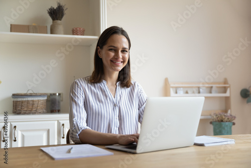 Beautiful young 25s woman sit at table in cozy home office, distracted from telecommute work on laptop smile look at camera. Portrait of happy freelancer, comfort remote online services user concept