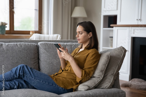 Side view young serious woman sit on sofa holds smartphone texting messages, share sms make distance communication with friend on-line. Weekend leisure use modern wireless tech, mobile apps concept photo