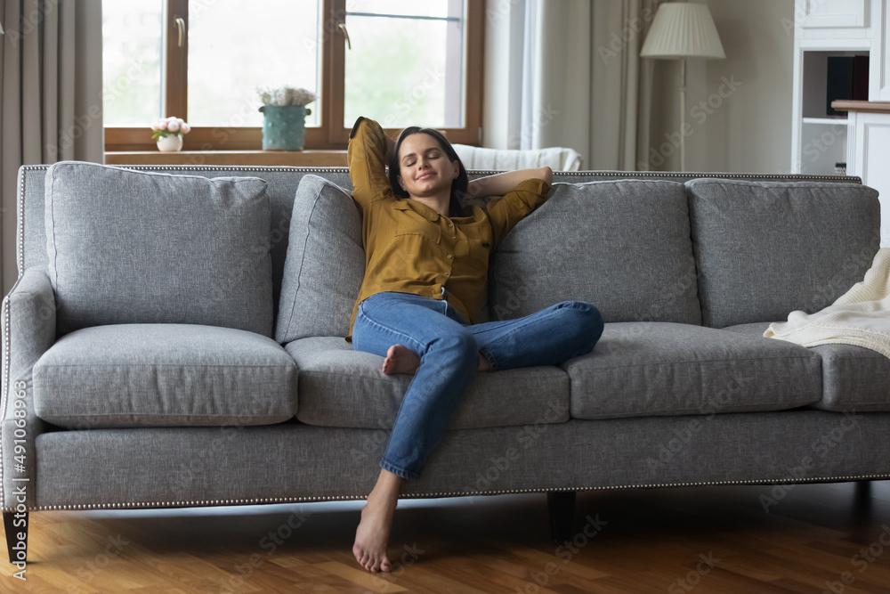 Peaceful attractive smiling young woman stretching on cozy couch with hands behind head closed her eyes breath fresh conditioned air inside. Weekend at home, modern smart house owner, tenant concept Peaceful attractive smiling young woman stretching on cozy couch with hands behind head closed her eyes breath fresh conditioned air inside. Weekend at home, modern smart house owner, tenant concept