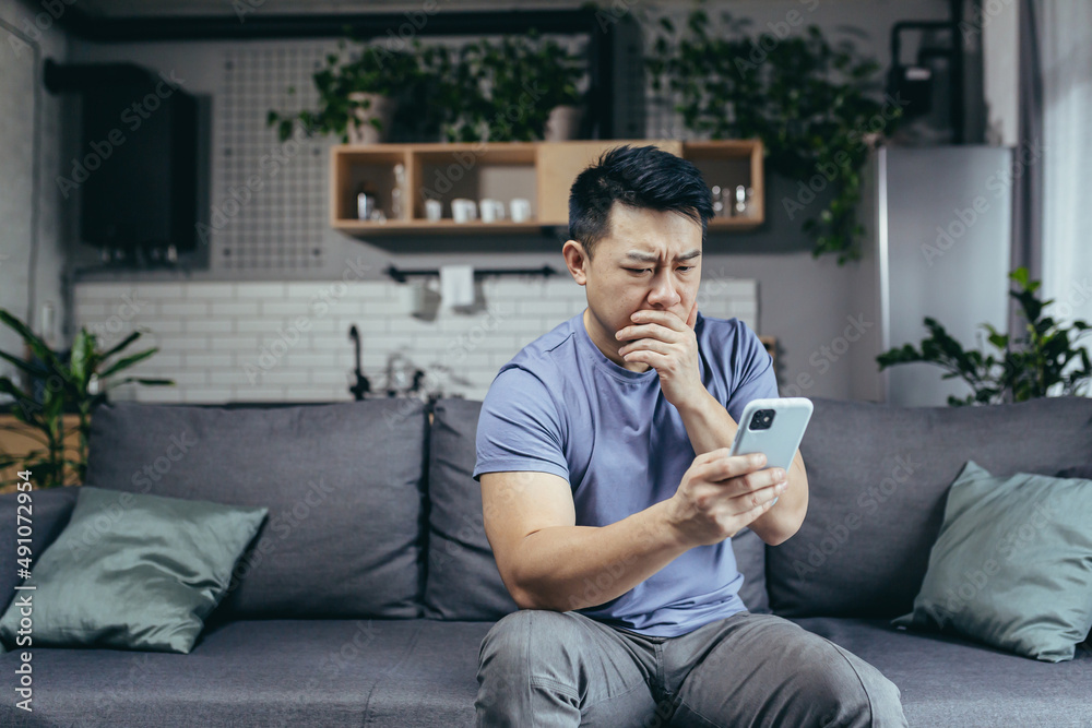 Man reading bad news from phone, asian frustrated and sad looking at phone screen sitting on sofa at home Man reading bad news from phone, asian frustrated and sad looking at phone screen sitting on sofa at home