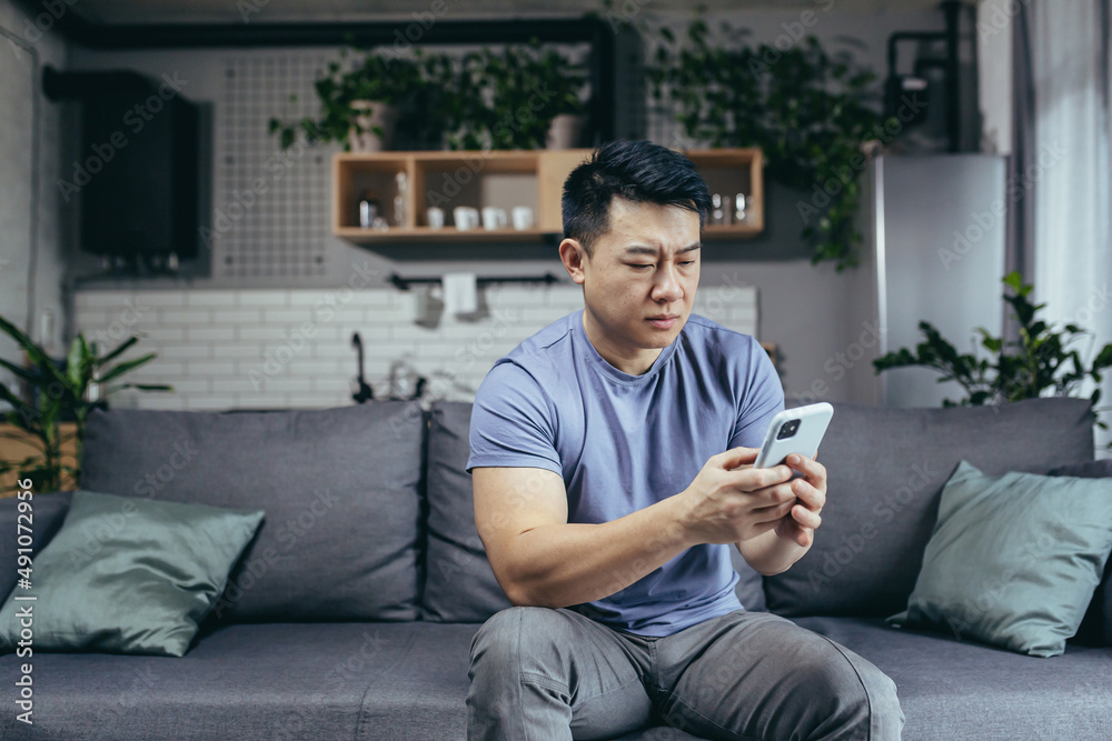 Man reading bad news from phone, asian frustrated and sad looking at phone screen sitting on sofa at home