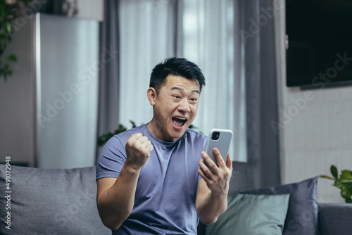 Happy Asian at home, won in the online uses the application on the smartphone, the man shows a gesture of victory, happy and satisfied sitting on the couch