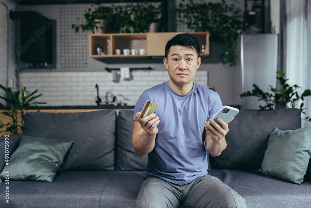 Fototapeta premium man alone at home, shopping online, asian uses phone and shopping app, holds credit card, sits on sofa at home