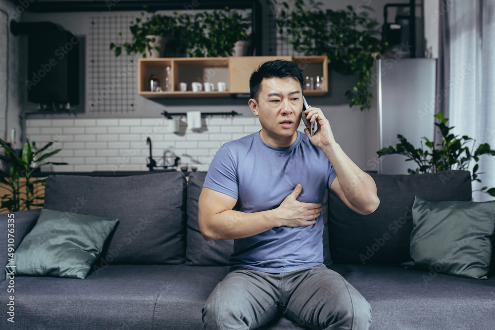 Asian man at home alone, has severe chest pain, calls a doctor, reports his illness, sitting on the couch at home