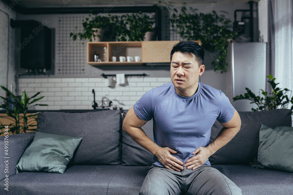 Asian man himself at home sick sitting on the couch, has severe abdominal pain, man in the living room Asian man himself at home sick sitting on the couch, has severe abdominal pain, man in the living room