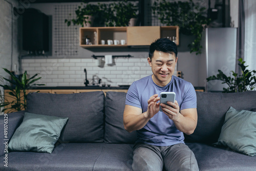 Happy asian at home sitting on sofa enjoying phone, typing message, cheerful and smiling man in loft living room