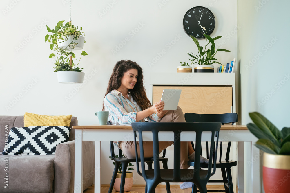Cheerful smiling business woman talking on a video call meeting on her digital tablet while drink cup of coffee and sitting at home office desk Cheerful smiling business woman talking on a video call meeting on her digital tablet while drink cup of coffee and sitting at home office desk