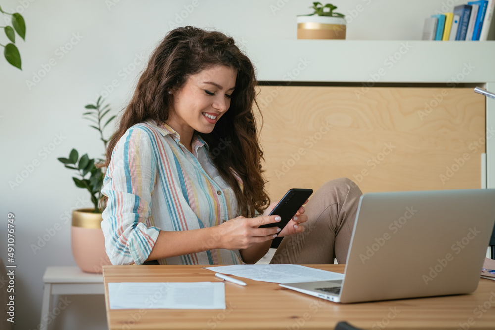 Happy Businesswoman Using Mobile Phone at Home Office. 
Cheerful smiling business woman reading or typing text message on her phone while sitting at desk and working on her laptop computer and papers.