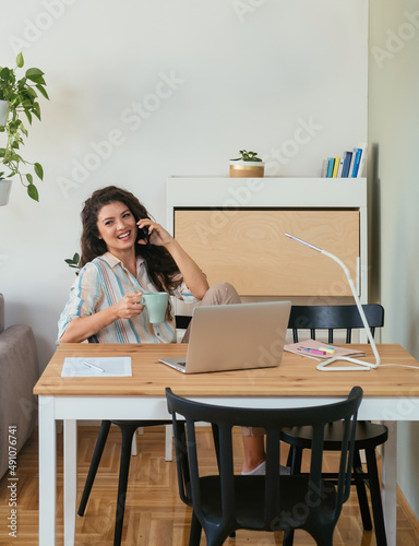 Happy Businesswoman Talking on a Mobile Phone at Home. Cheerful smiling business woman speaking on her phone while holding a cup of coffee and working on her laptop computer.