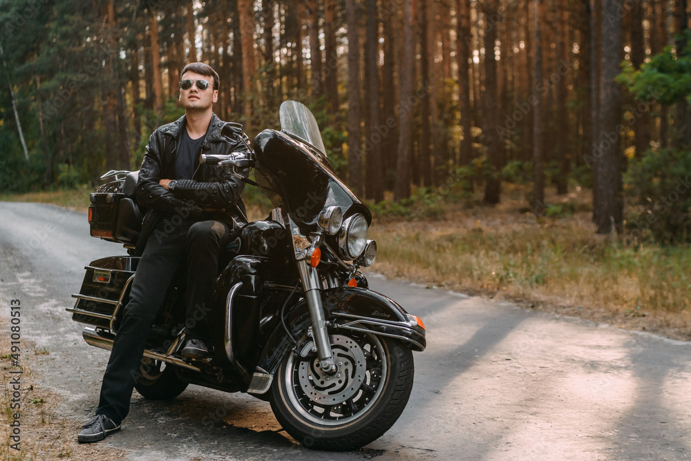A biker man in a leather jacket and sunglasses is sitting on his motorcycle.
