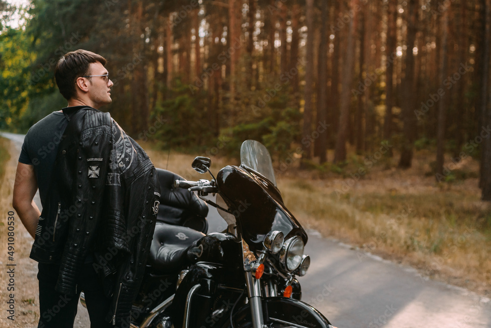Selective focus of young man in black leather jacket standing near motorcycle