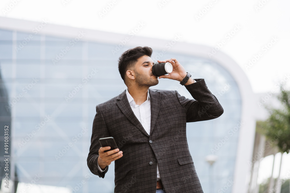 Young handsome Indian businessman standing in front of modern building or airport, smiling and talking on cell phone, drinking take away coffee Young handsome Indian businessman standing in front of modern building or airport, smiling and talking on cell phone, drinking take away coffee