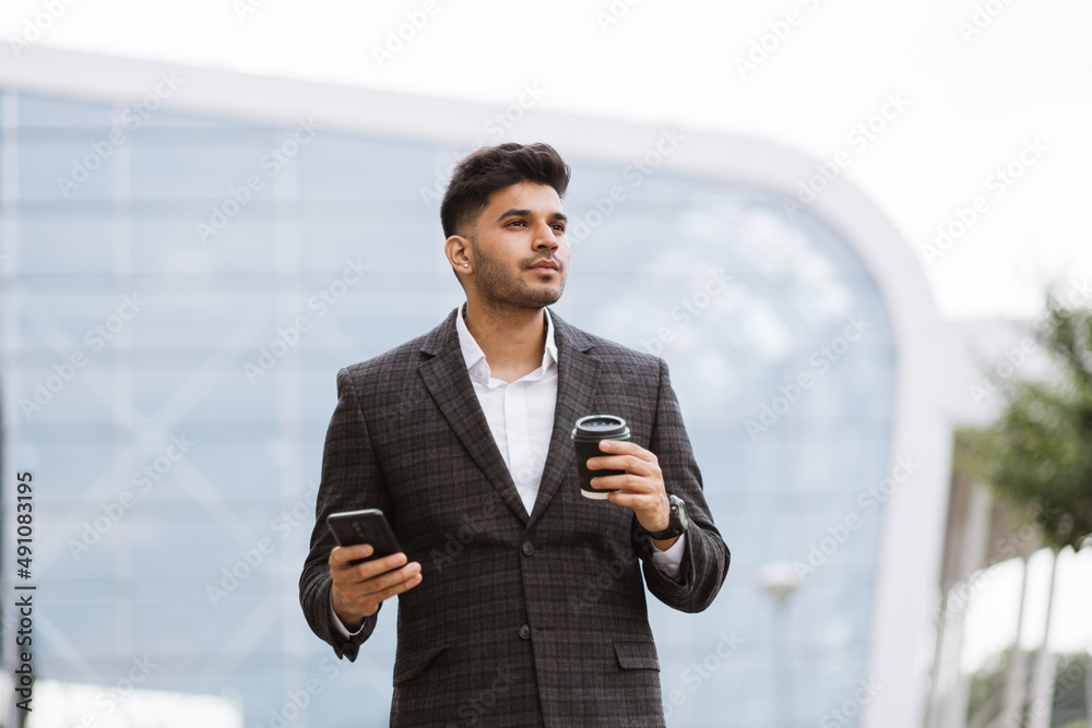 Handsome Indian businessman in formal wear, holding a cup of coffee looking away. Young thoughtful Arab man standing outside modern building and drinking take away coffee Handsome Indian businessman in formal wear, holding a cup of coffee looking away. Young thoughtful Arab man standing outside modern building and drinking take away coffee