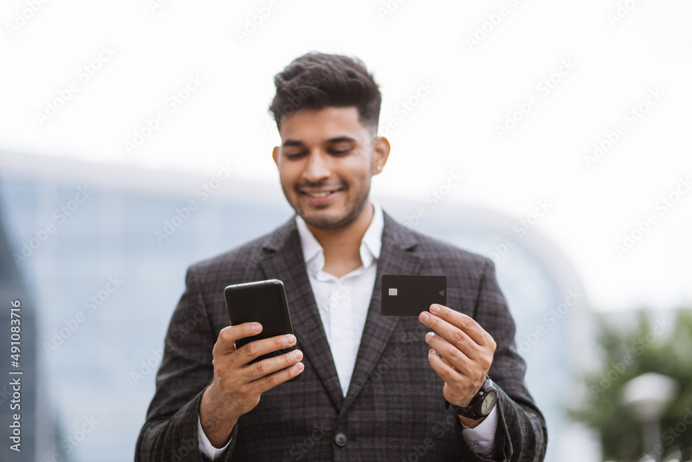 Business trip concept. Handsome young Indian man in eyeglasses, using smart phone and holding credit card with shopping online. Male freelancer receives payment online Business trip concept. Handsome young Indian man in eyeglasses, using smart phone and holding credit card with shopping online. Male freelancer receives payment online