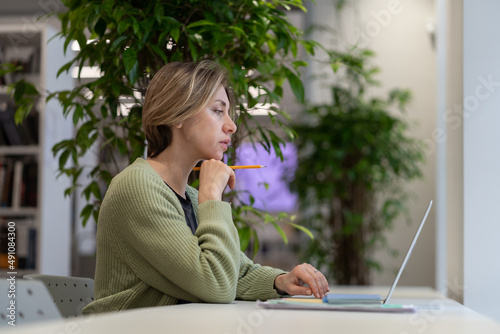 Focused pensive woman university professor preparing lecture material on laptop in light cozy library with green houseplants, selective focus. Scandinavial middle-aged female getting second degree photo