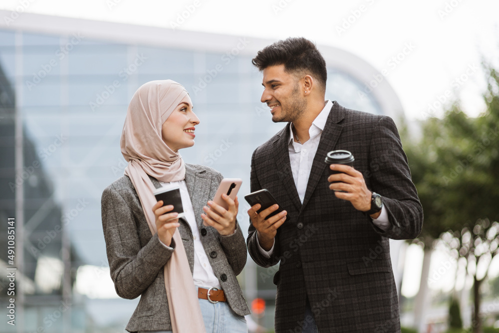 Arabian business couple having fun and talking together walking on the street during coffee break. Woman in hijab showing her smartphone screen to her colleague, sharing idea or news