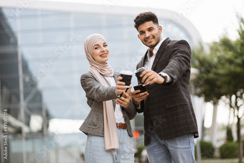 Smiling business couple, handsome Arab man and Muslim woman in hijab, standing outdoors in the street on coffee break, using phones and drinking take away coffee © sofiko14