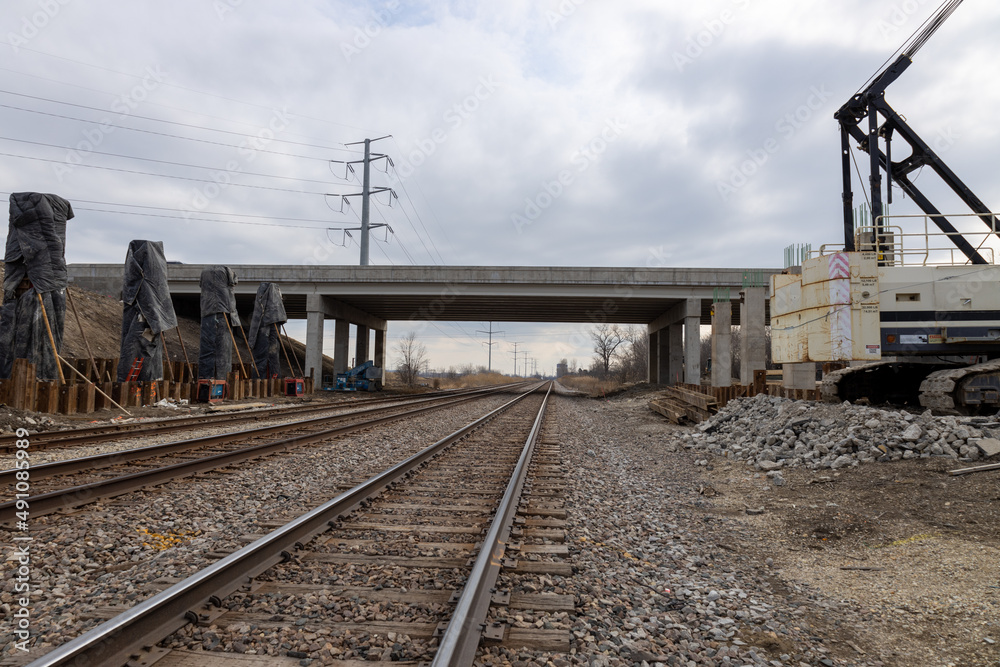 View of newly constructed highway overpass over train track. Crane on one side to help with construction. Piling with newly poured concrete covered from the weather. Dark gray clouds in sky. View of newly constructed highway overpass over train track. Crane on one side to help with construction. Piling with newly poured concrete covered from the weather. Dark gray clouds in sky.