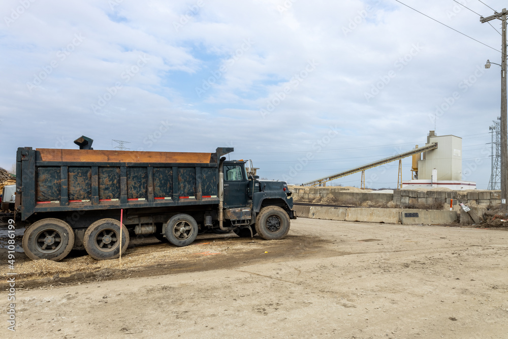 Fototapeta premium Green dump truck parked in yard awaiting its next trip. Blue sky with fluffy clouds. Concrete barrier wall. Gravel elevator for taking rock and sand to make concrete. Transformer towers seen in back. 