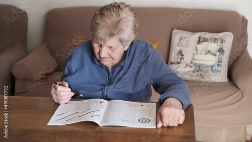 Elderly woman solves crossword puzzle. Senior woman sitting on a sofa with a comforter on her legs playing crossword. Brain and memory training. Enhancing short term memory skills.
