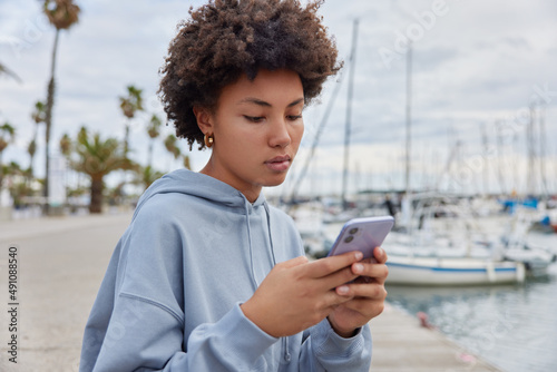 Serious curly haired woman wears blue sweatshirt uses mobile phone surfs social media checks notifications poses against sea harbor dressed in casual hoodie. People and modern lifestyle concept
