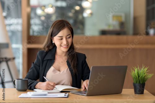 Beautiful young Asian businesswoman is smiling at her desk and taking notes with computer laptop on her desk, enjoying work.