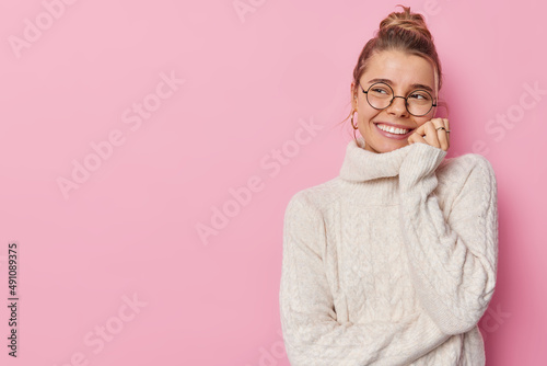 Horizontal shot of pretty young woman with hair bun smiles gently looks away with glad expression listens something pleasant wears spectacles and knitted sweater poses indoor against pink wall