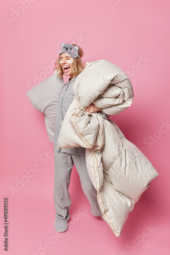 Overjoyed young woman laughs gladfully dressed in pajama feels very glad holds soft duvet and pillow undergoes beauty procedures after sleeping isolated over pink background. Good night concept