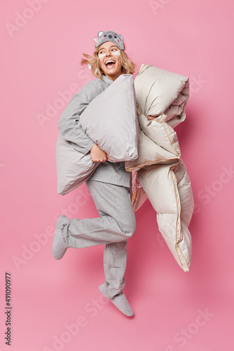 Vertical shot of happy woman dressed in pajama holds pillow and blanket wakes up early in morning applies beauty patches under eyes looks gladfully away isolated over pink backgroud. Bedtime concept
