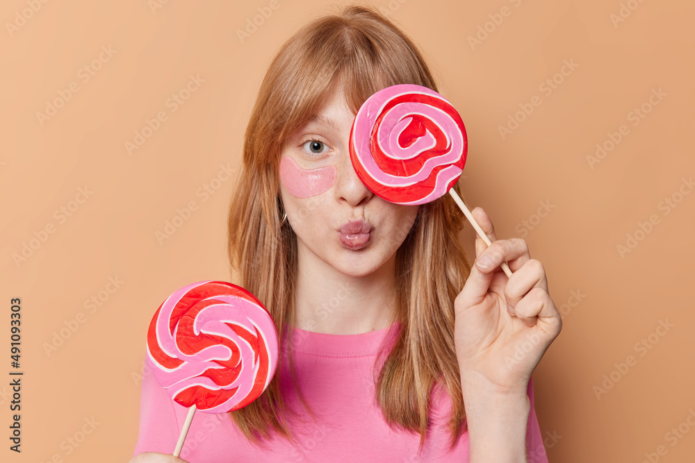 Horizontal shot of redhead girl covers eye with round caramel candy keeps lips folded applies hydrogel patches under eyes for skin treatment enjoys eating sweets isolated over beige background
