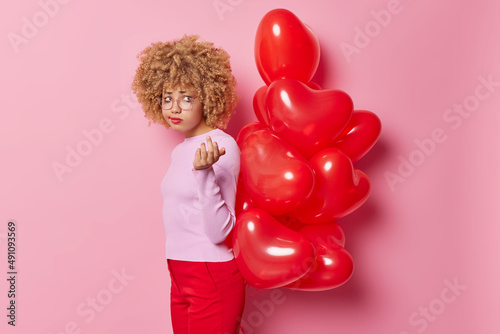 Frustrated curly haired young woman has sad expression looks unhappily somewhere holds bunch of heart balloons behind back wears casual clothes prepares surprise isolated over pink background