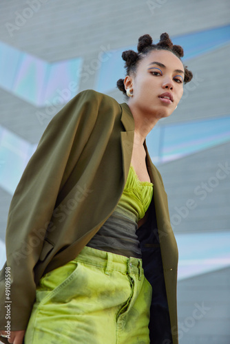 Fashionable woman with trendy hairstyle dressed in green fashionable clothes looks confidently at camera wears blue eyeliner poses against blurred background. View from top. People and style