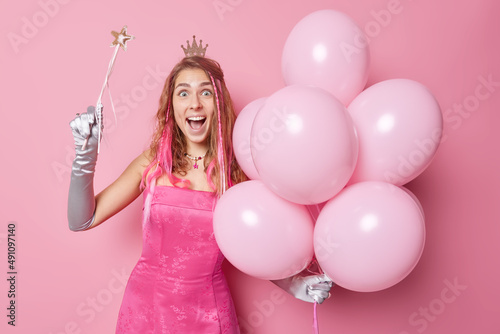 People special occasion and entertainment concept. Positive young pretty woman princess wears crown on head dress long gloves makes your wishes come true holds magic wand bunch of helium balloons photo