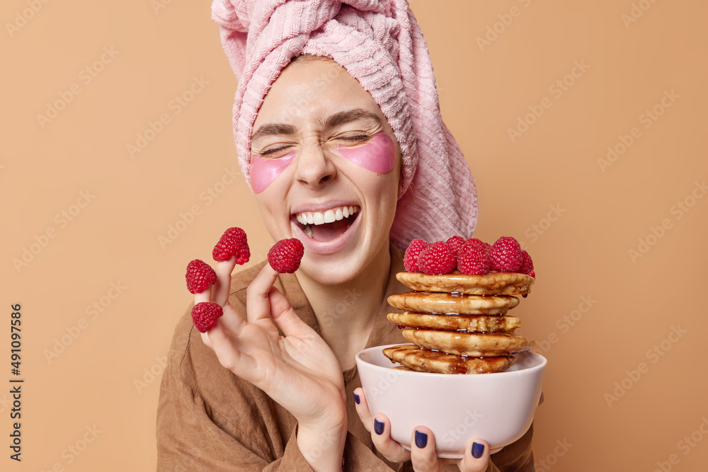 Morning time and delicious breakfast. Glad joyful woman holds delicious appetizing pancakes with raspberry has overjoyed expression wears bath towel on head and pajama isolated over brown wall Morning time and delicious breakfast. Glad joyful woman holds delicious appetizing pancakes with raspberry has overjoyed expression wears bath towel on head and pajama isolated over brown wall