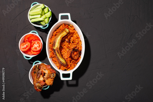 Jollof rice with fried banana, baked chicken. Rice with tomatoes, onions, spices. Traditional national Nigerian food. Dark background. View from above. Place for text. photo