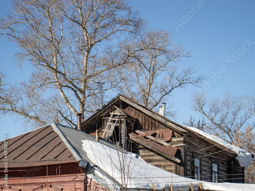 View of the roofs of old houses under the snow. Snow lies on the roofs of houses. Roofs of houses and trees without leaves against a blue sky without clouds. © Vladimir