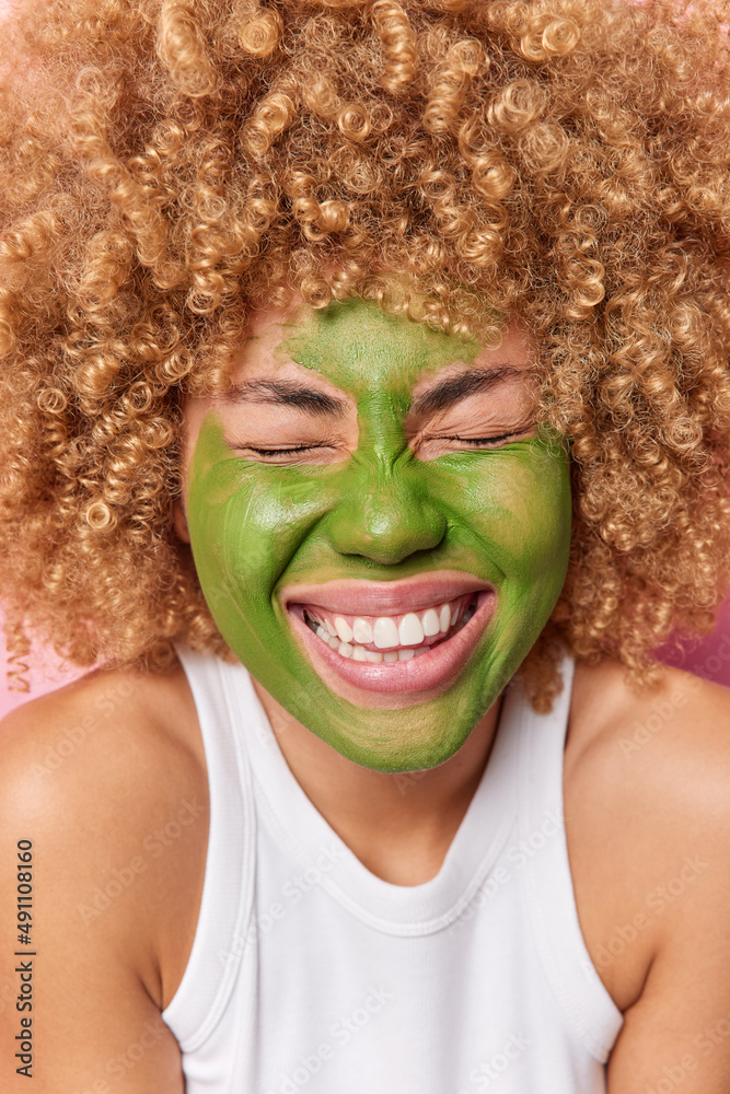 Vertical shot of happy curly haired woman closes eyes smiles broadly shows white teeth applies green beauty mask for skin treatment wears casual white t shirt. Wellness facial care and spa concept Vertical shot of happy curly haired woman closes eyes smiles broadly shows white teeth applies green beauty mask for skin treatment wears casual white t shirt. Wellness facial care and spa concept