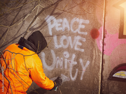 a guy in a hood draws graffiti Peace, love, unity on the wall.