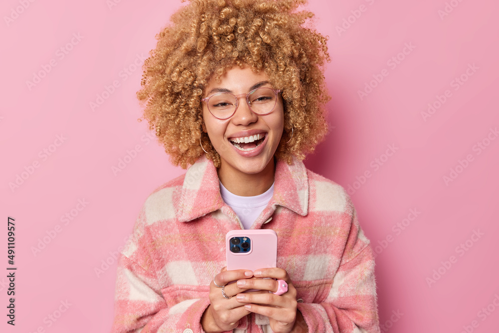 Indoor shot of happy curly haired woman laughs joyfully dressed in casual checkered jacket uses mobile phone connected to wifi types feedback enjoys cellphone messaging isolated over pink background