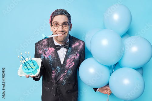 Indoor shot of happy brunet birthday man blows party horn holds festive cake and bunch of inflated balloons dressed in formal suit isolated over blue background meets guests. Enternaiment concept photo