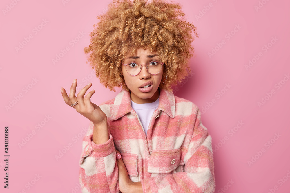 Indignant woman with curly hair raises hand and shrugs shoulders purses lips wears checkered jacket has hesitant expression isolated over pink background. So what should I do in this situation