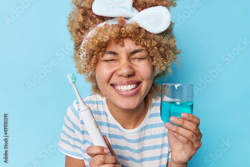 Oral care concept. Positive young curly woman smiles toothily keeps eyes closed holds electric brush and mouthwash dressed casually undergoes daily hygiene routines poses against blue background
